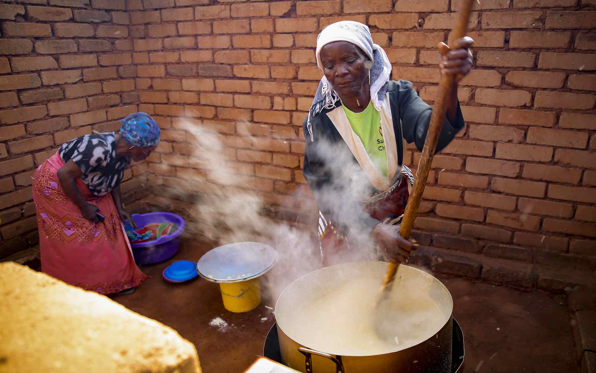 Woman, center, stirs porridge in a big pot; woman, left, reaches into a plastic tub