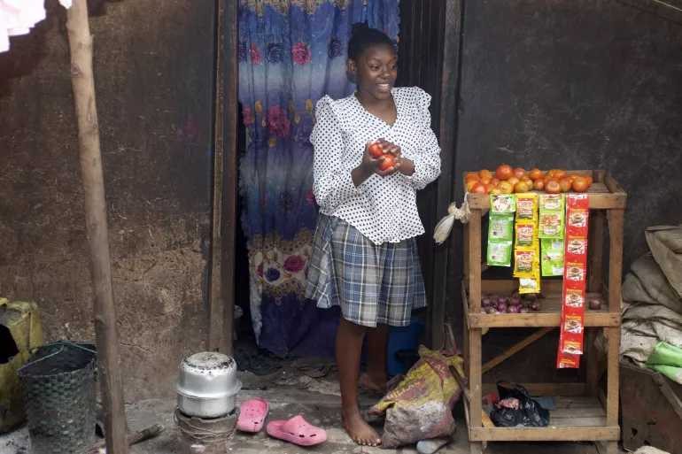 Young woman standing in doorway holding tomatoes next to cart with tomatoes, other items for sale