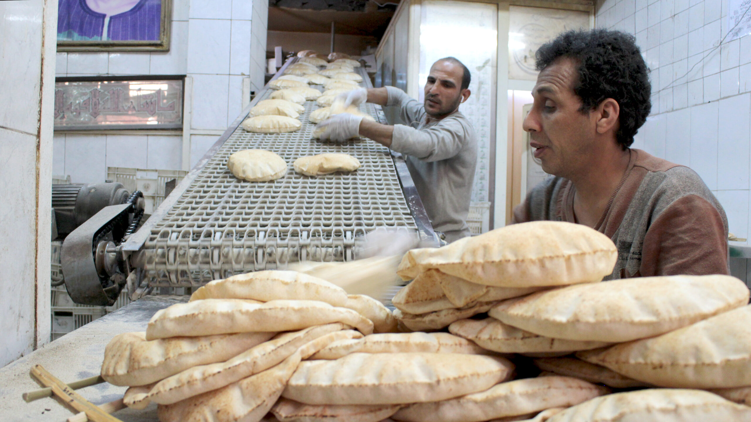 Pile of bread in foreground, man behind, man further back next to ramp emerging from overn with loaves rolling down.