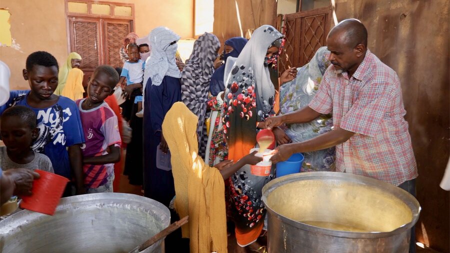 Man, right, behind large cauldrons, pours stew into girl's cup; women lined up behind them