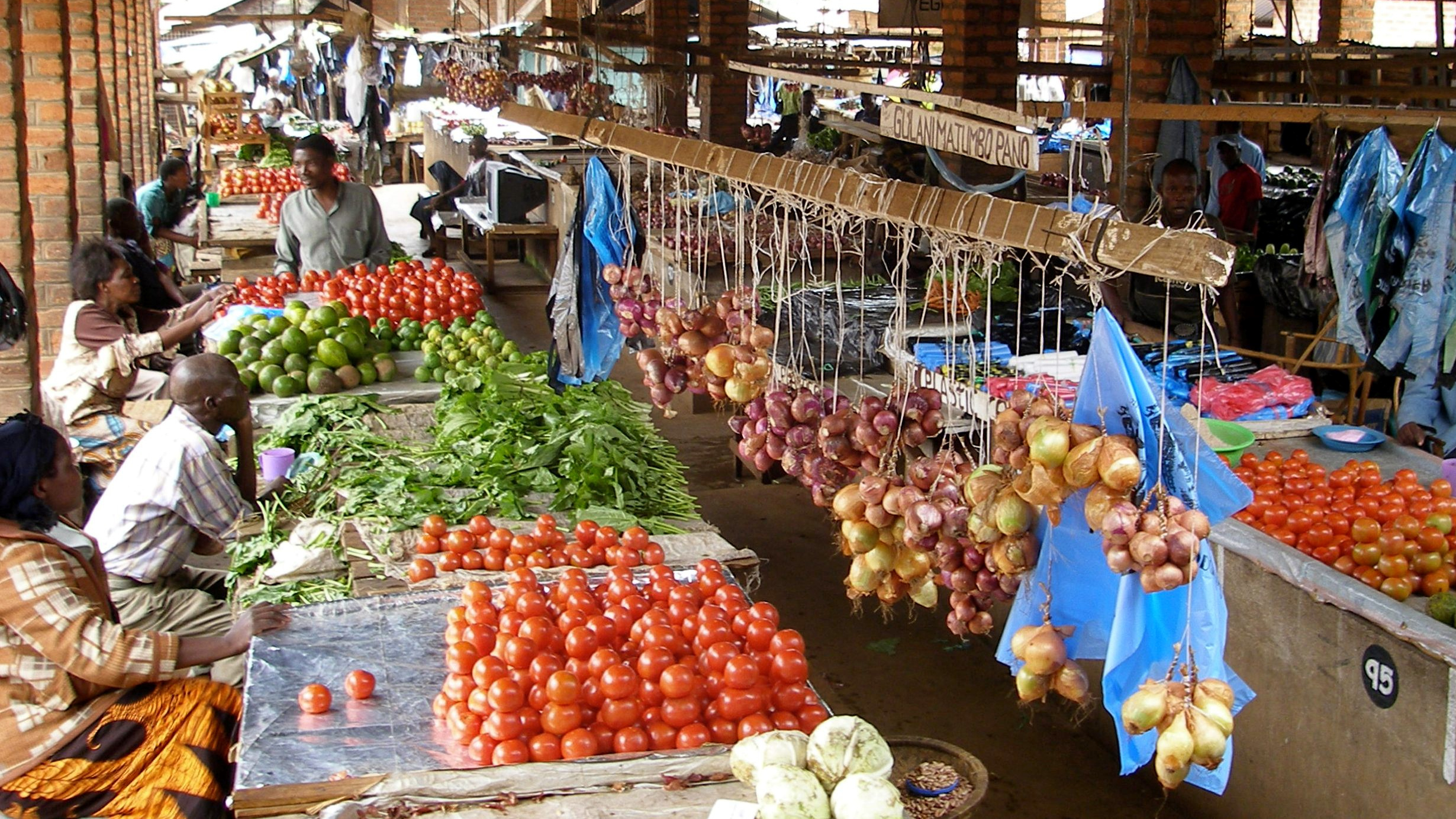 Vendors, seated on left, behind tables of tomatoes, greens, and hanging bunches of onions in market