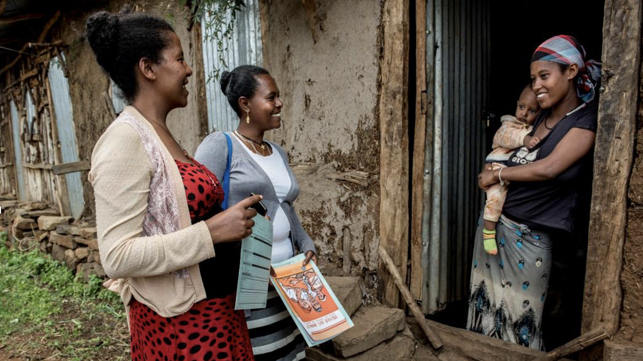 Two women, left, one holding printed materials, talking with woman, right, in doorway of house holding baby.