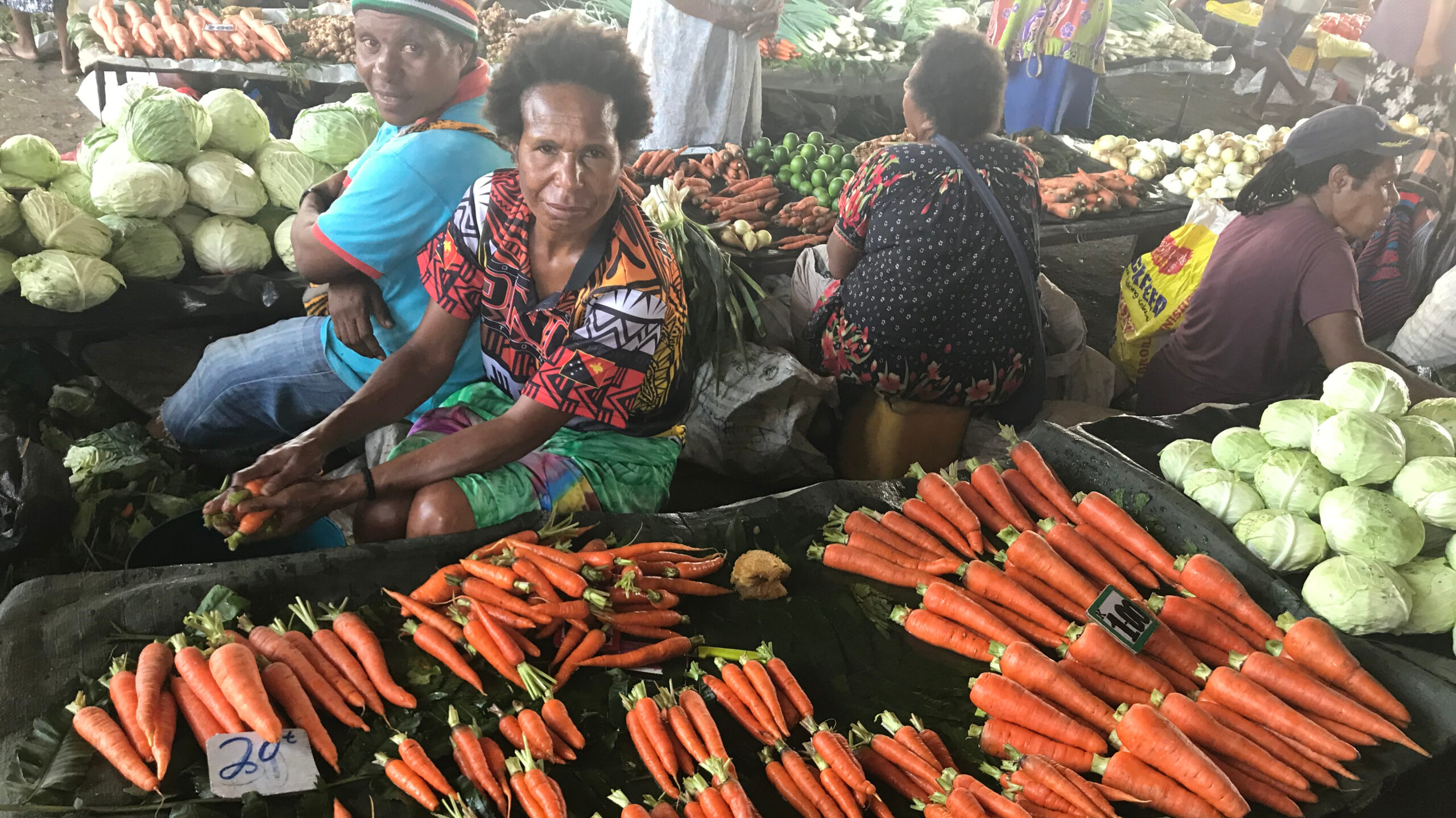 Four women vendors seated between rows of produce laid out for sale, carrots on table in foreground.