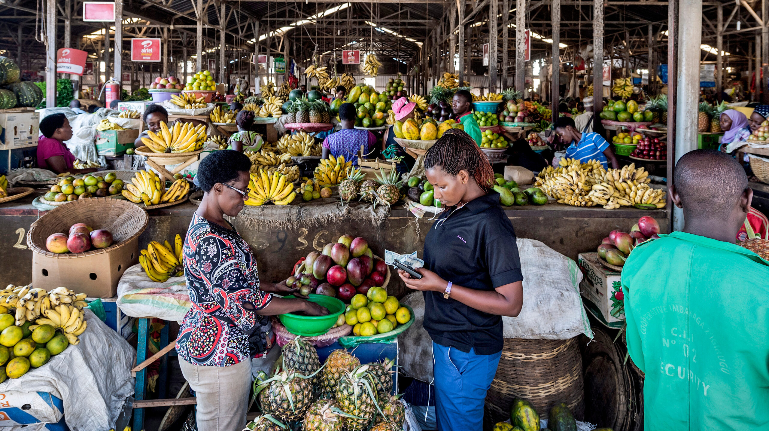 Two women facing each other, one holding money, foreground, in center of large covered produce market.