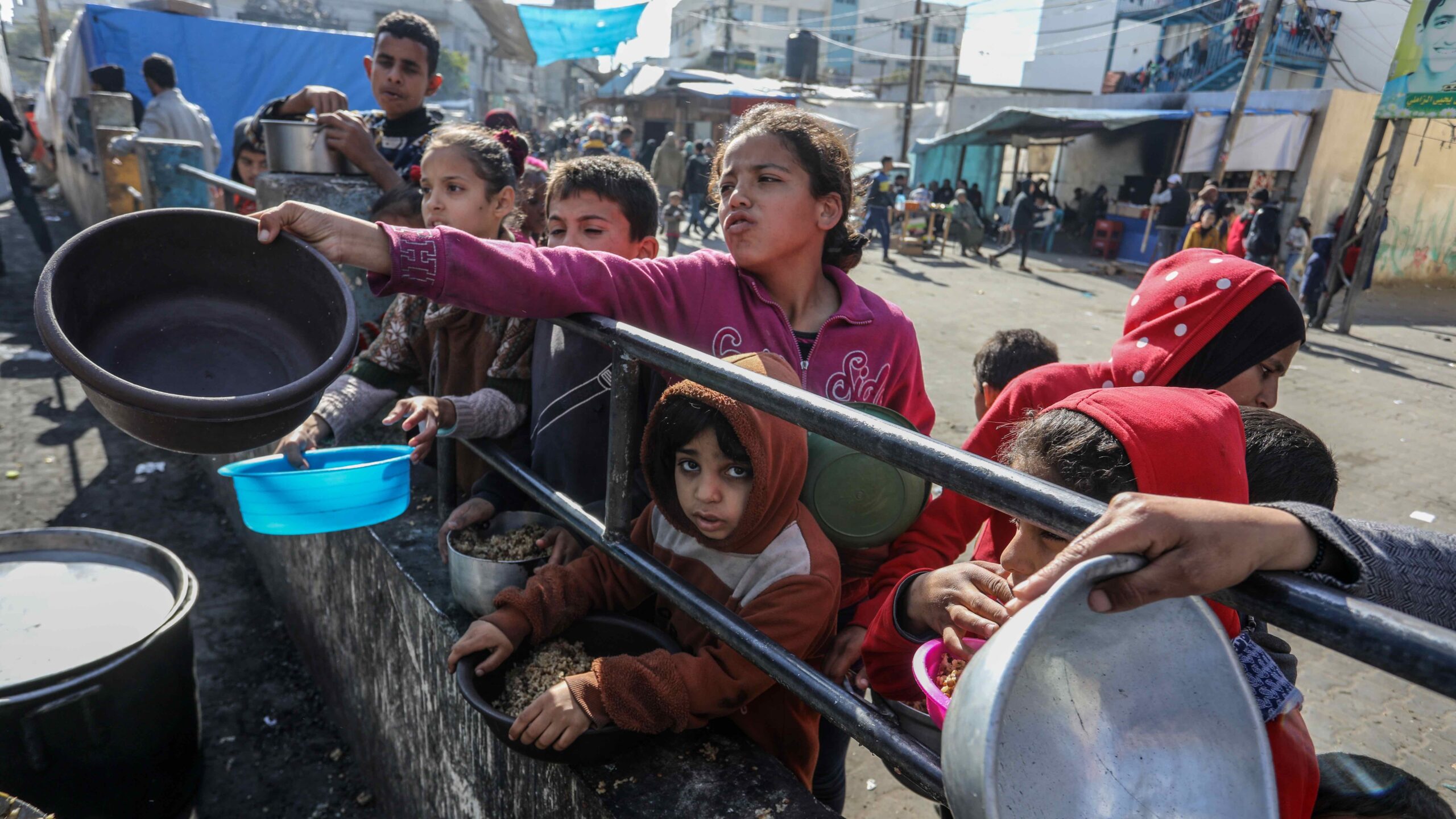 Children holding bowls and containers lined up along a railing. Girl, center, reaches her bowl across.