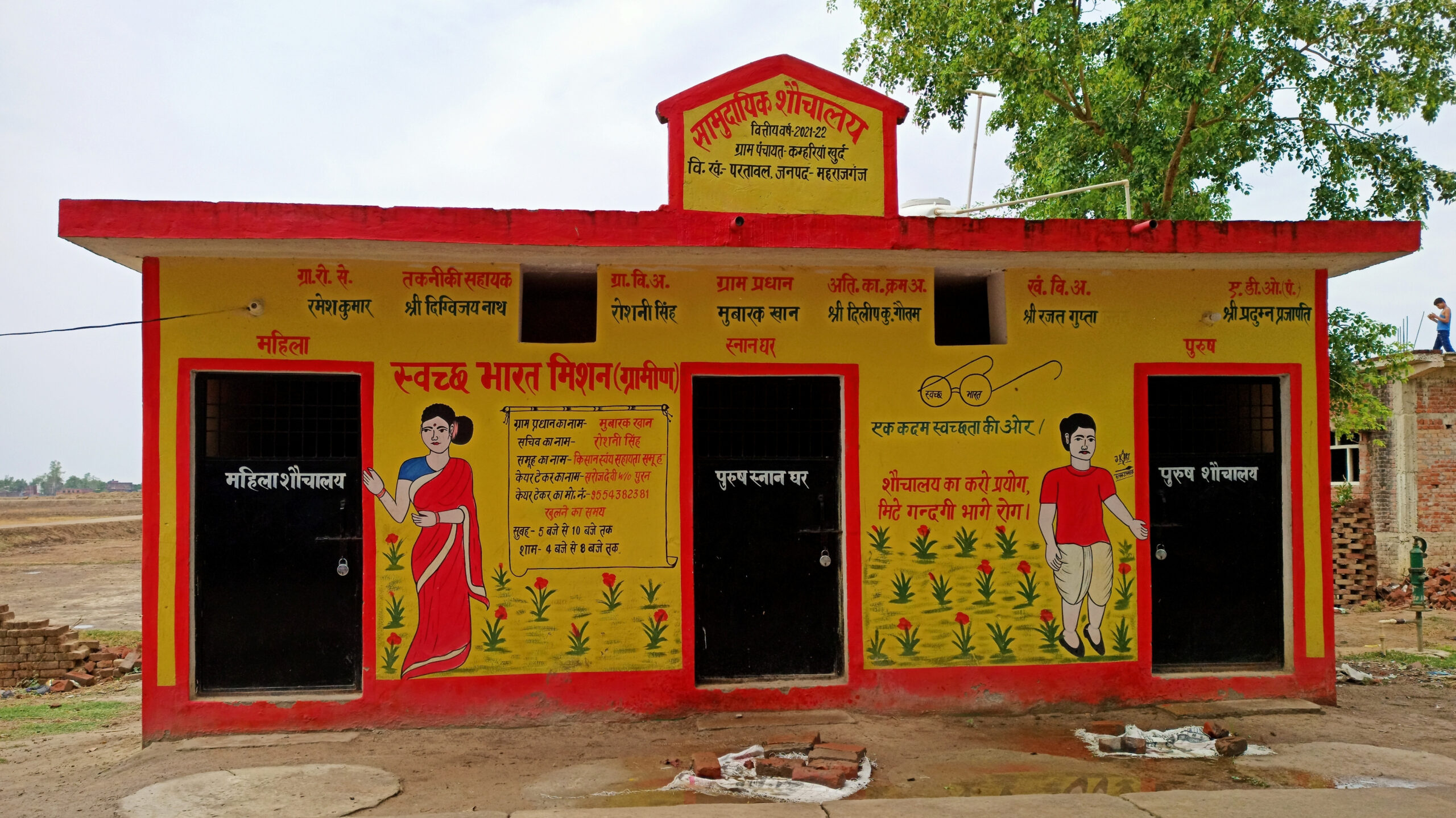 Public toilet building with three doors, painted yellow with red trim, with painted images of a man, woman, and plants and writing promoting the Swachh Bharat Mission.