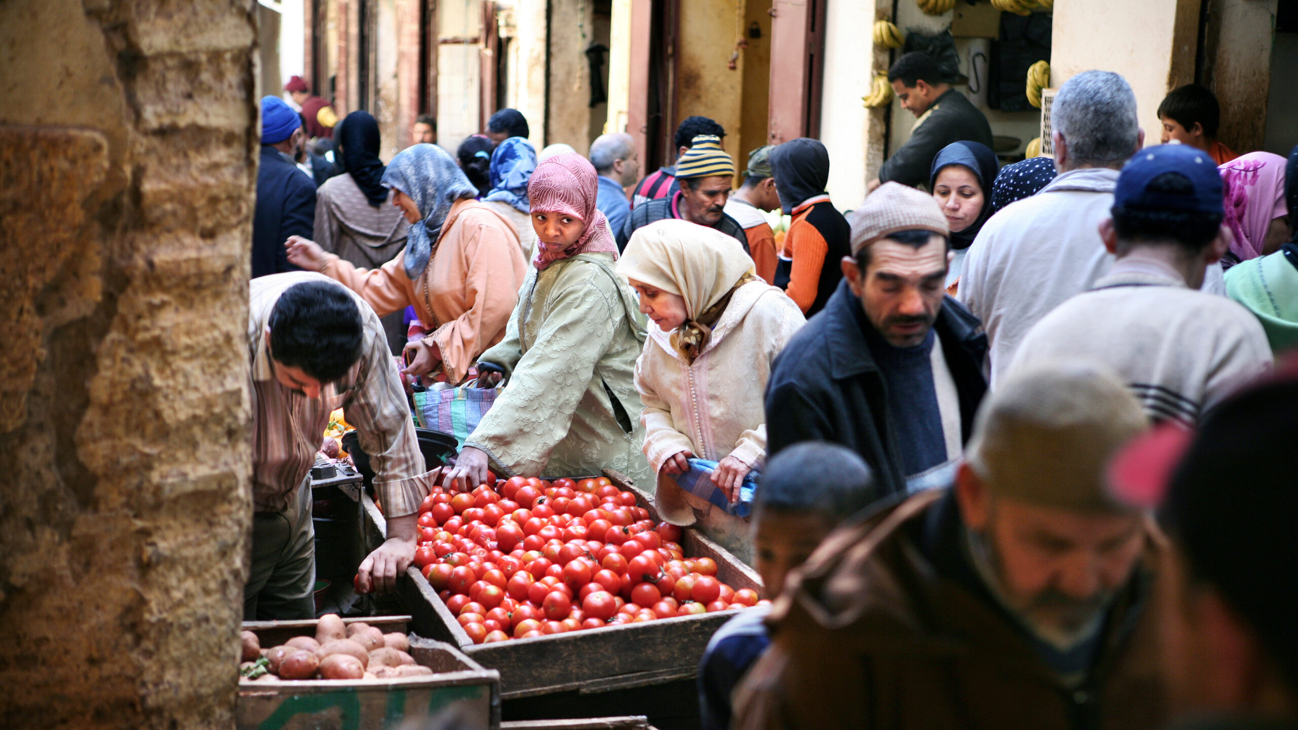 Two woman, center, wearing hijabs, look at a vat of tomatoes; vendor, a man, right, leans over. They are surrounded by people in a densely-packed street scene