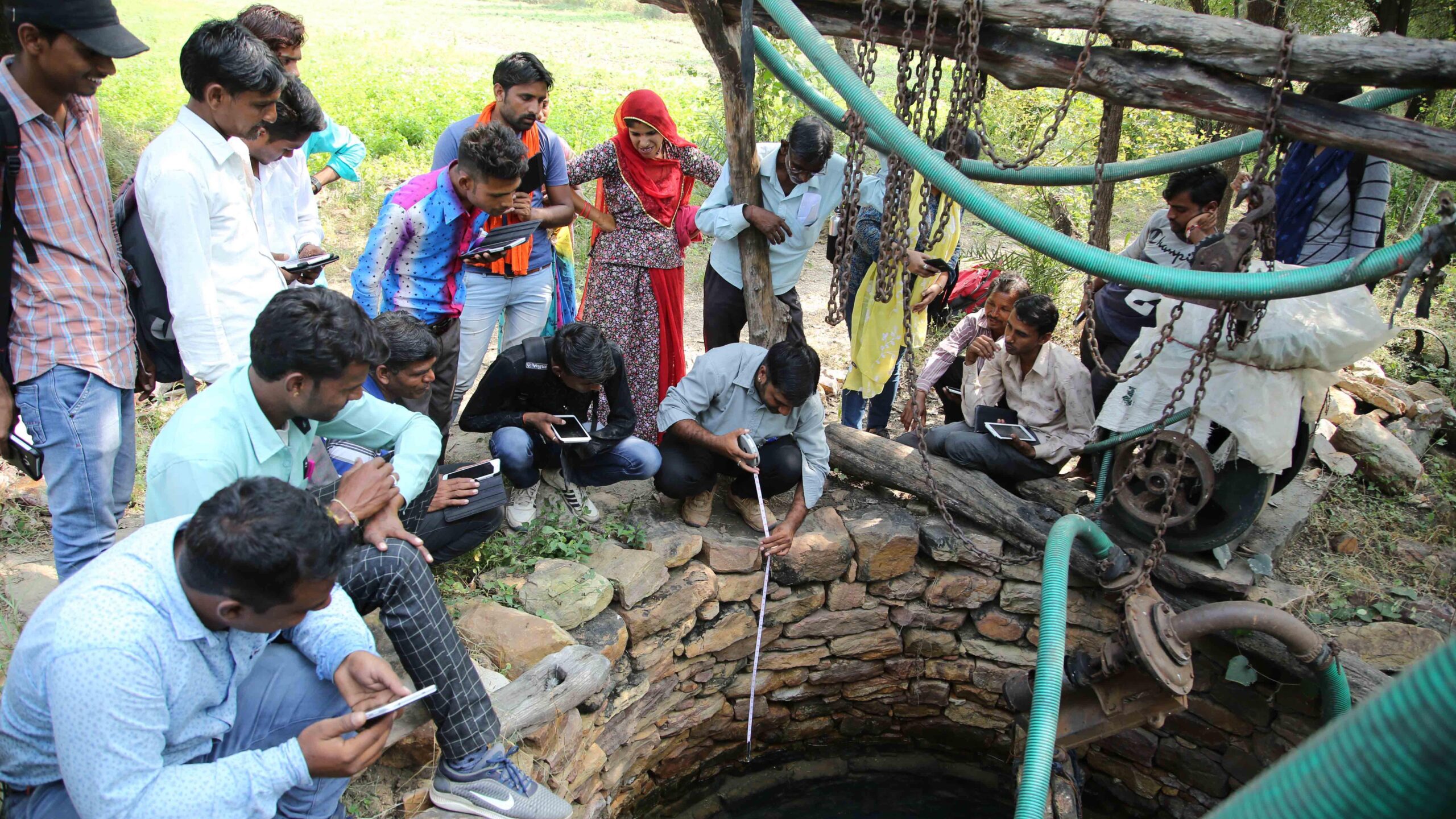 Group of men and women, many holding smartphones or tablets, around edge of a well; man, crounching center, holds measuring tape going down into well.