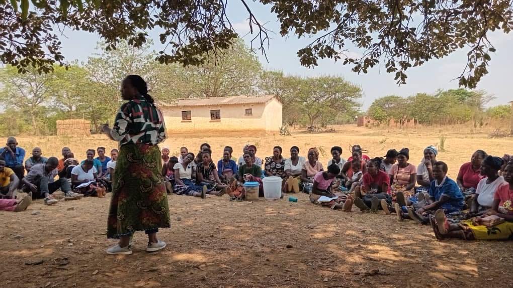 Woman, standing, foreground, talks to group seated on the ground in in semicircle around her