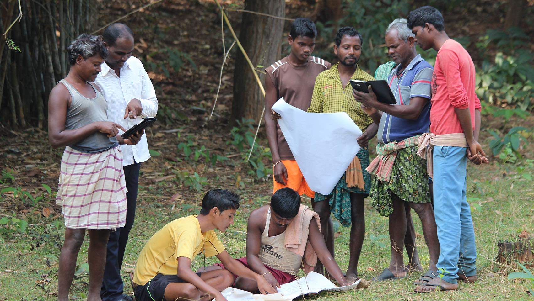 Four men, standing right examine tablet; one holds white paper; man and woman, standing left, examine table; two men sitting on ground between them examine large piece of paper