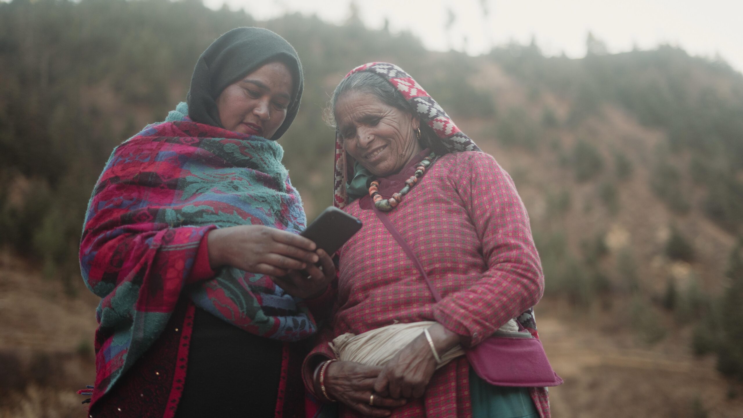 Two women standing outside look at smartphone held by woman on left.