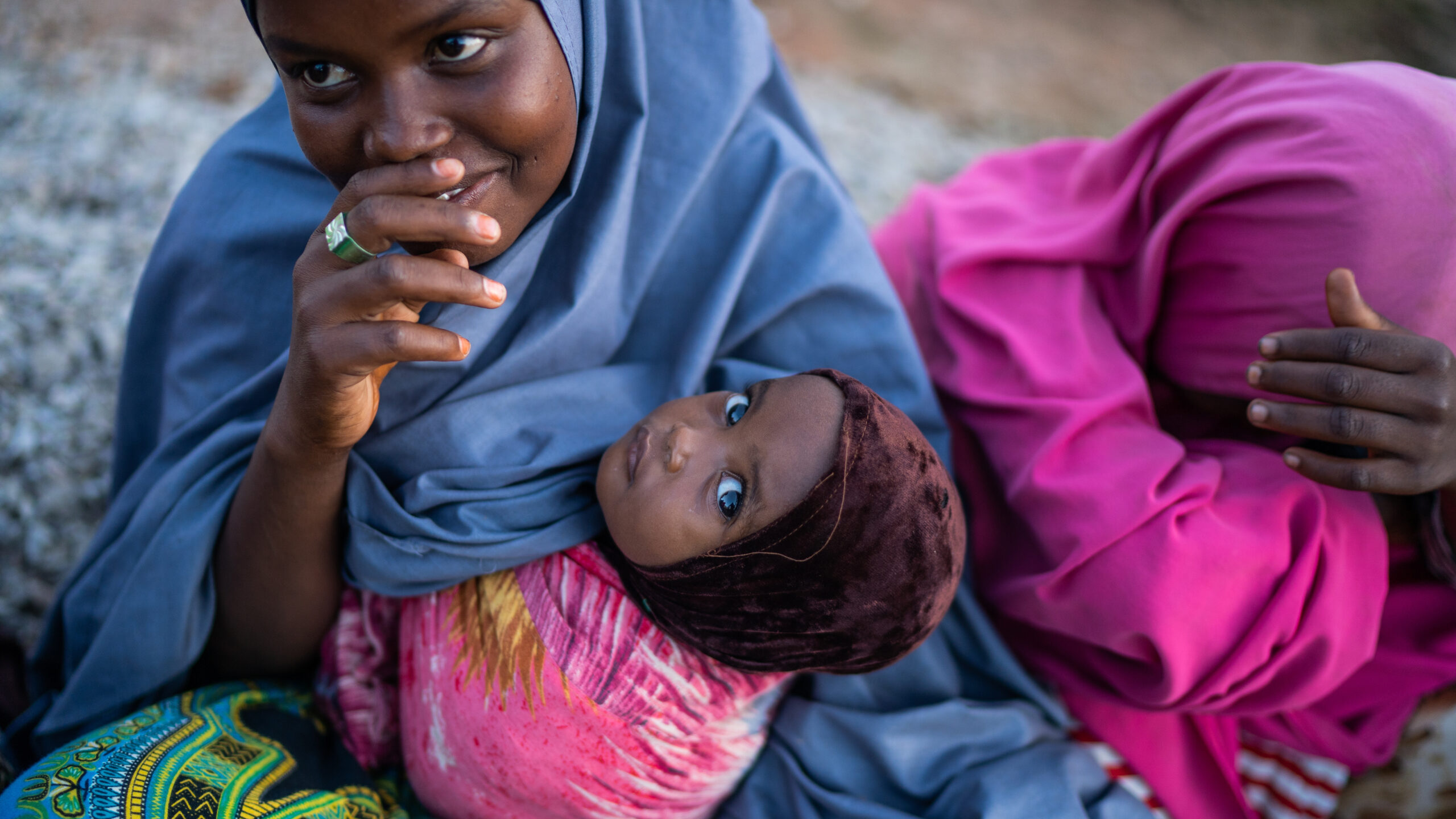 Mother, left, holds baby, seated on ground next woman, right