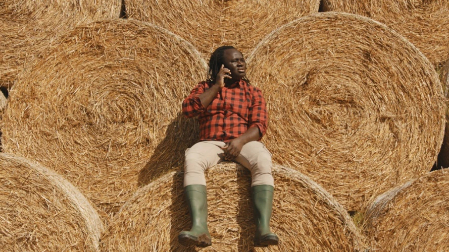 Man wearing boots sitting on round hay bale holding phone with more bales in around him