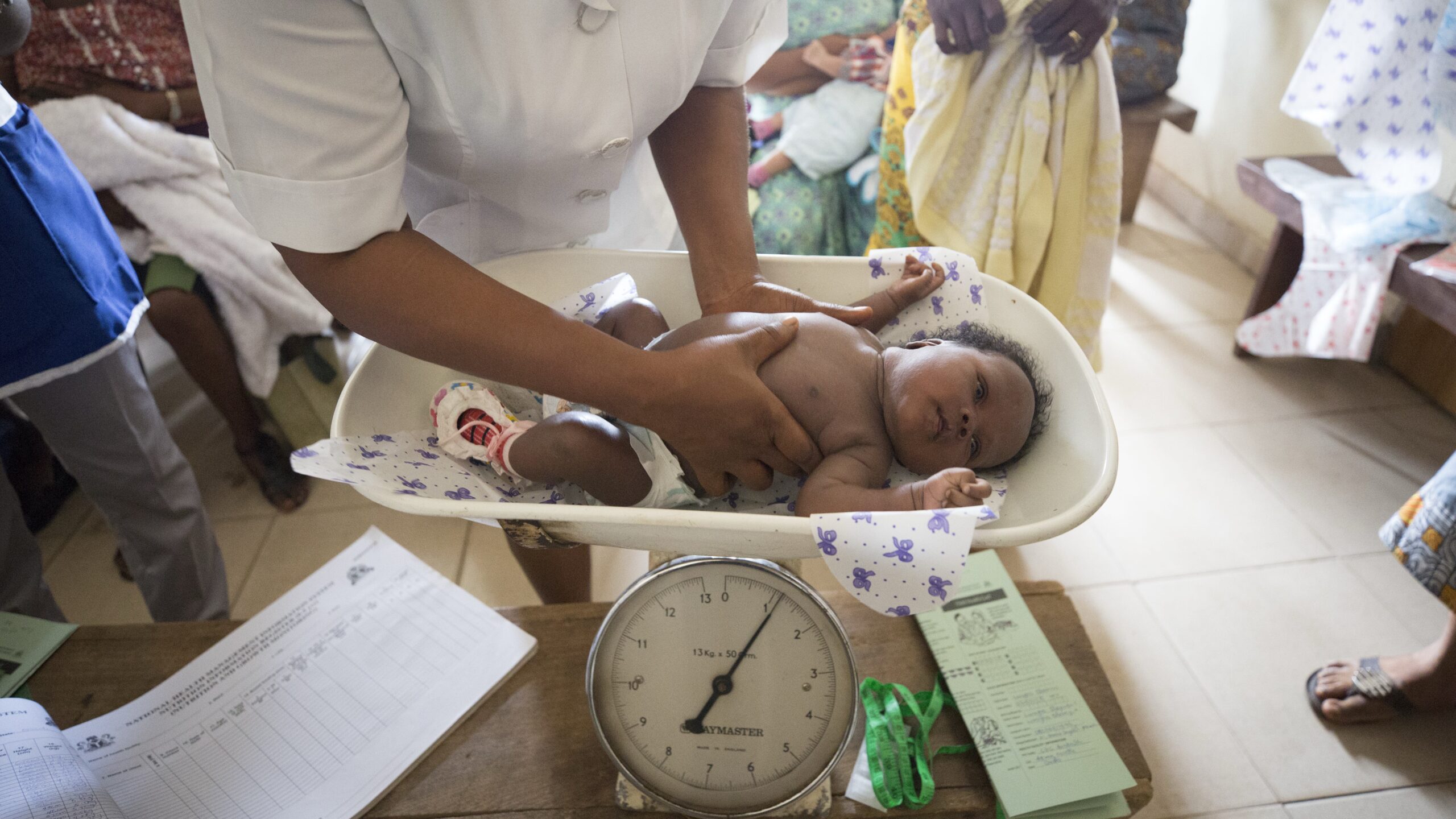 Baby being placed on scale by woman, other figures behind and to the right.